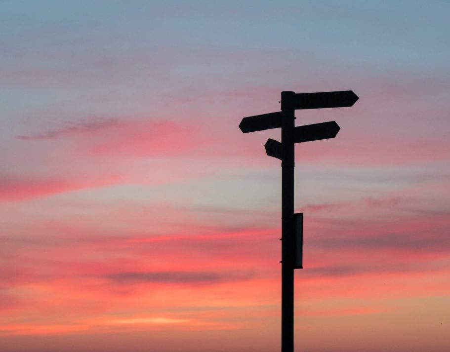 Symbolfoto Wegweiser Wegweiser mit mehreren Richtungspfeilen vor einem Sonnenuntergang in Pastelltönen.