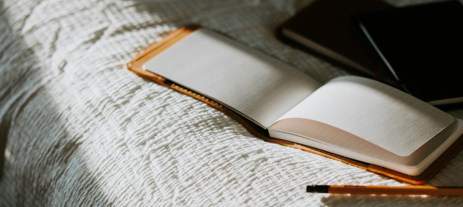 Symbolfoto Reporterblock Offenes Notizbuch auf einer weißen Decke, daneben einige Stifte und ein Notebook.